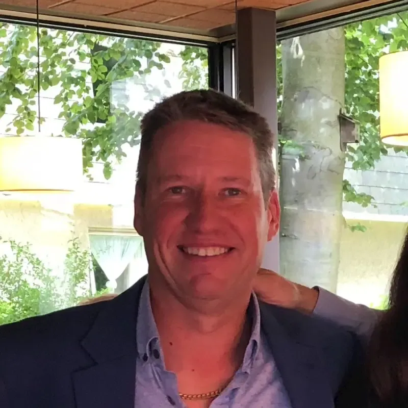 A smiling man with graying hair wearing a blue blazer and gold chain, with a window and trees visible in the background.