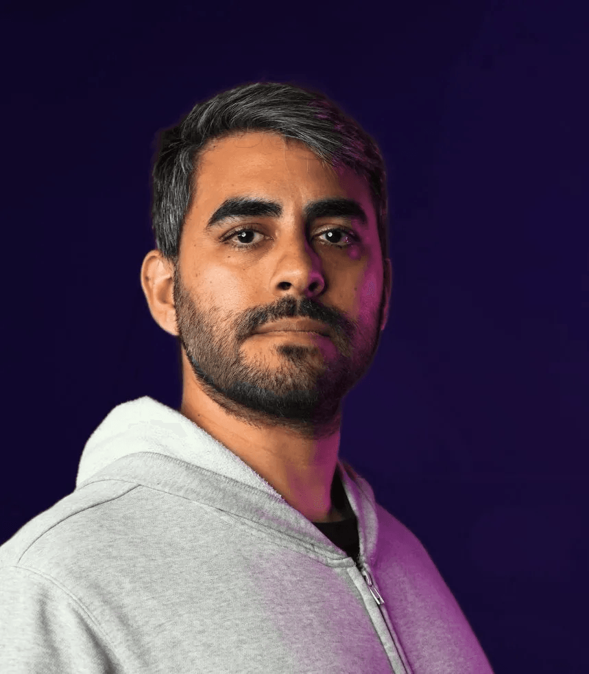 A man with a beard and salt-and-pepper hair, wearing a grey zippered hoodie, gazes directly at the viewer with a serious expression against a dark purple background with magenta lighting.