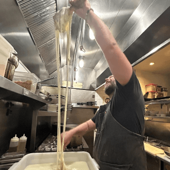 A chef in a professional kitchen stretches long strings of fresh white cheese upwards from a large tub, intently observing the process.