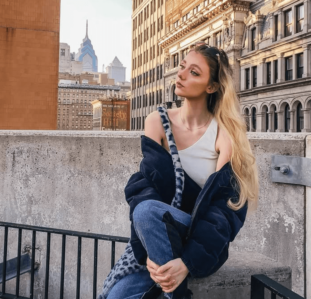 A young woman in a white tank top and blue jacket sits on a concrete ledge, looking thoughtfully at the Philadelphia city skyline with Society Hill.