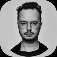 Black and white headshot of a man with dark curly hair, a beard, and round glasses, looking forward.