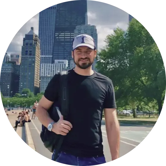 Smiling man in a black t-shirt and light baseball cap, carrying a backpack, stands on a city waterfront with tall buildings and people relaxing in the background.