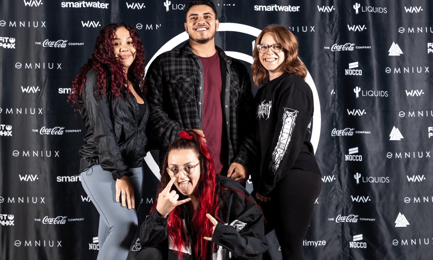 Four smiling young adults pose in front of a black event backdrop with sponsor logos, one with red hair making a rock-on gesture.