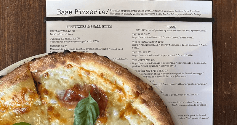 A close-up of a golden-crusted pizza with melted cheese, tomato, and fresh basil, with a Base Pizzeria menu highlighting local, organic ingredients.