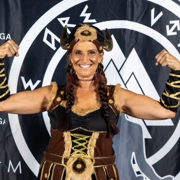 A smiling woman dressed in a Viking costume with a horned headpiece and braids flexes both biceps in front of a symbolic banner.