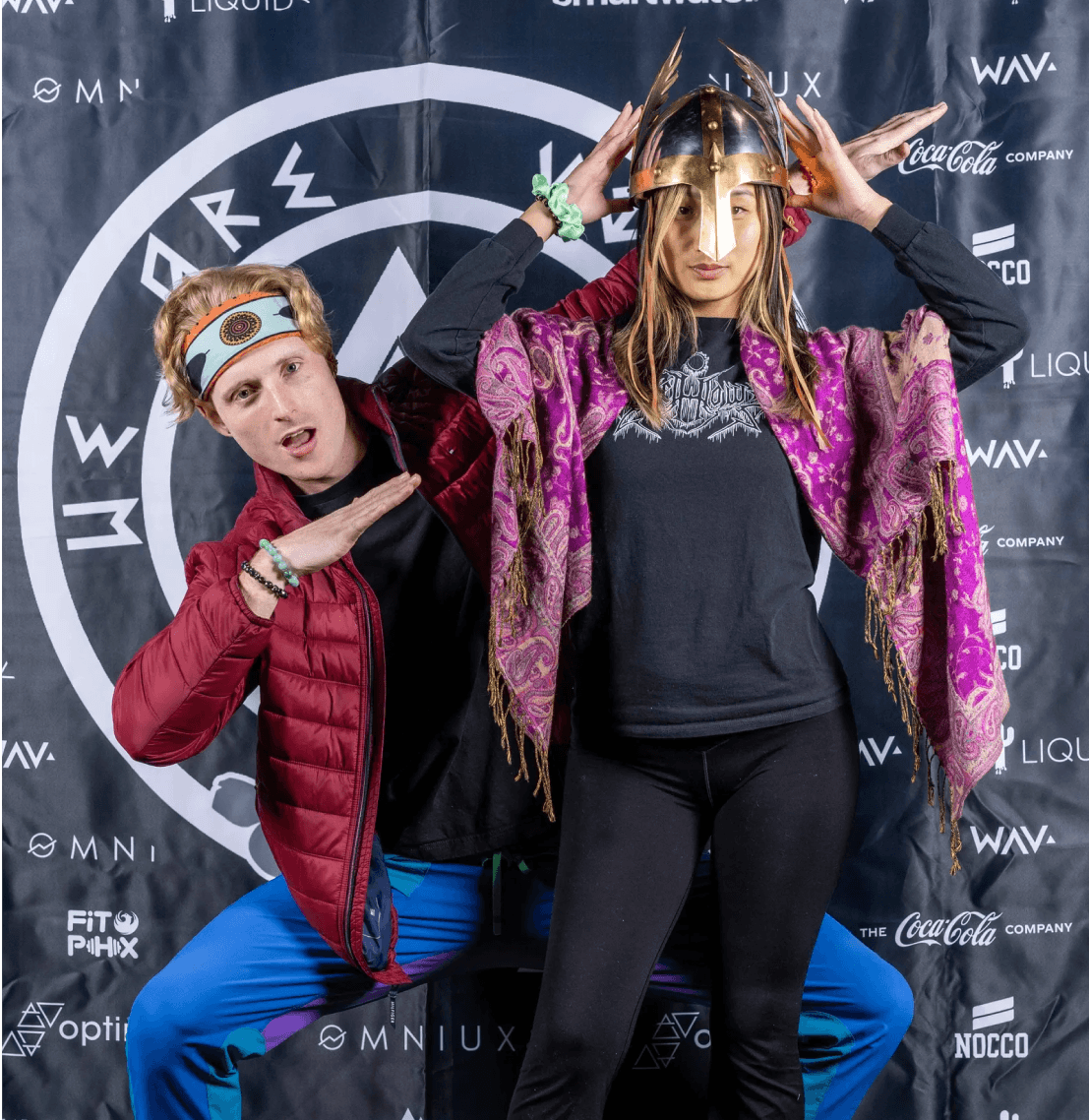 Two people pose playfully against a branded event backdrop; a man in a red jacket makes a dabbing gesture, and a woman wears a gold Viking helmet.